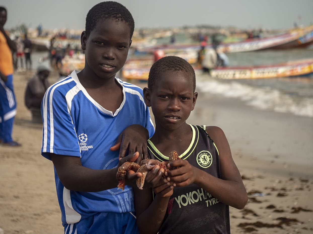 2 jeunes garçons tiennent des étoiles de mer à la plage au Sénégal, entourés de pêcheurs